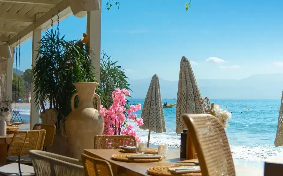 A shaded terrace looking out to the sea, cane chairs pulled up to a laid table and closed parasols on the sand.