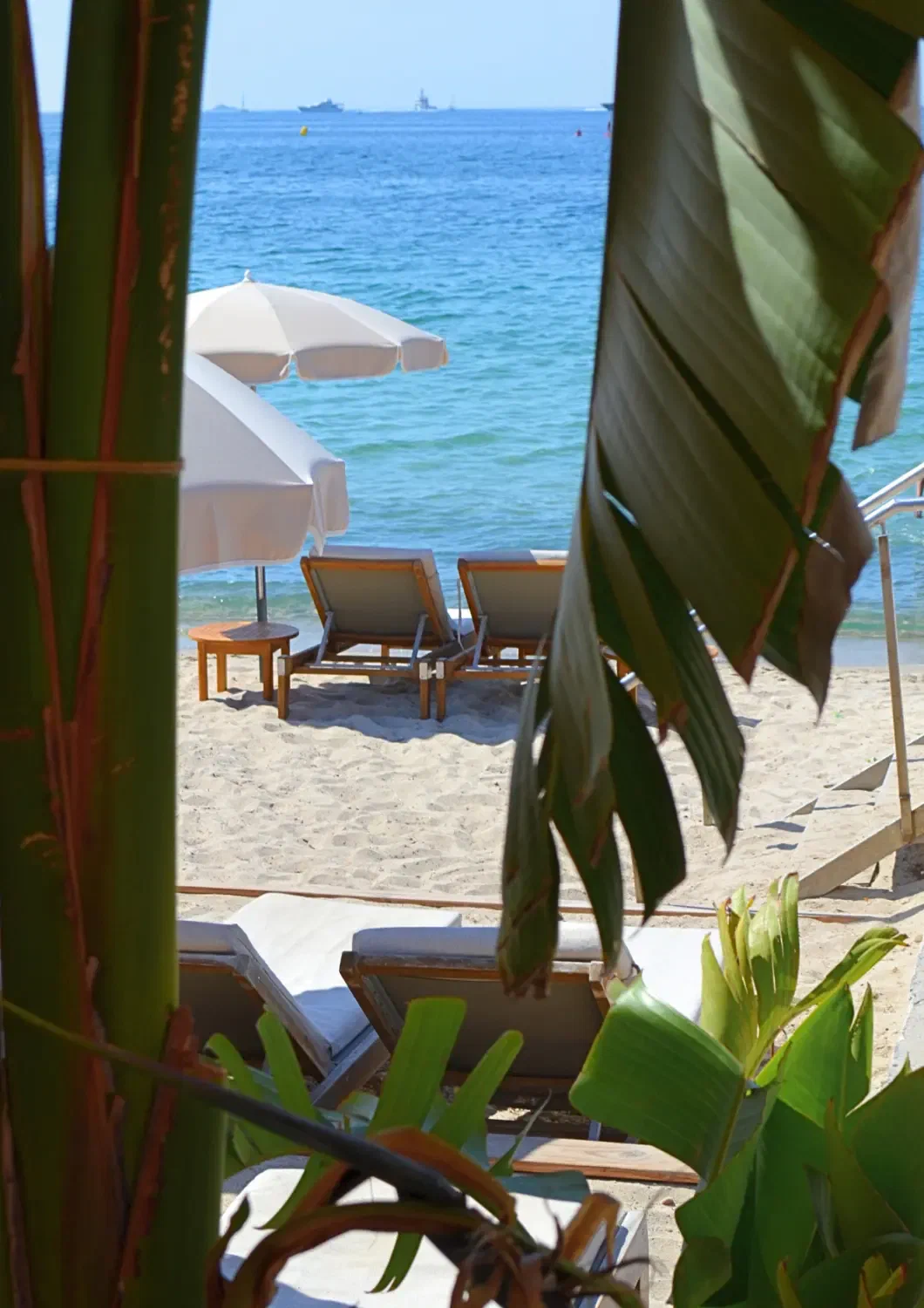 A gap between banana leaves opening onto two loungers and white parasols at the edge of the water.