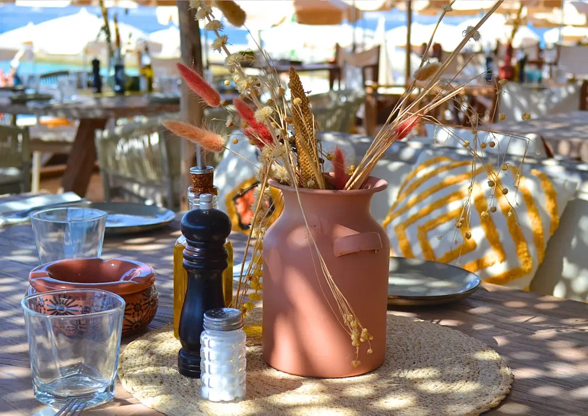 A terracotta jug of dried grasses on a wooden table, oil and glasses beside it in the sun.