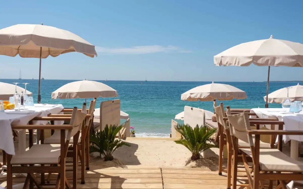 White-linen tables under cream parasols on either side of a sand path running down to the water.