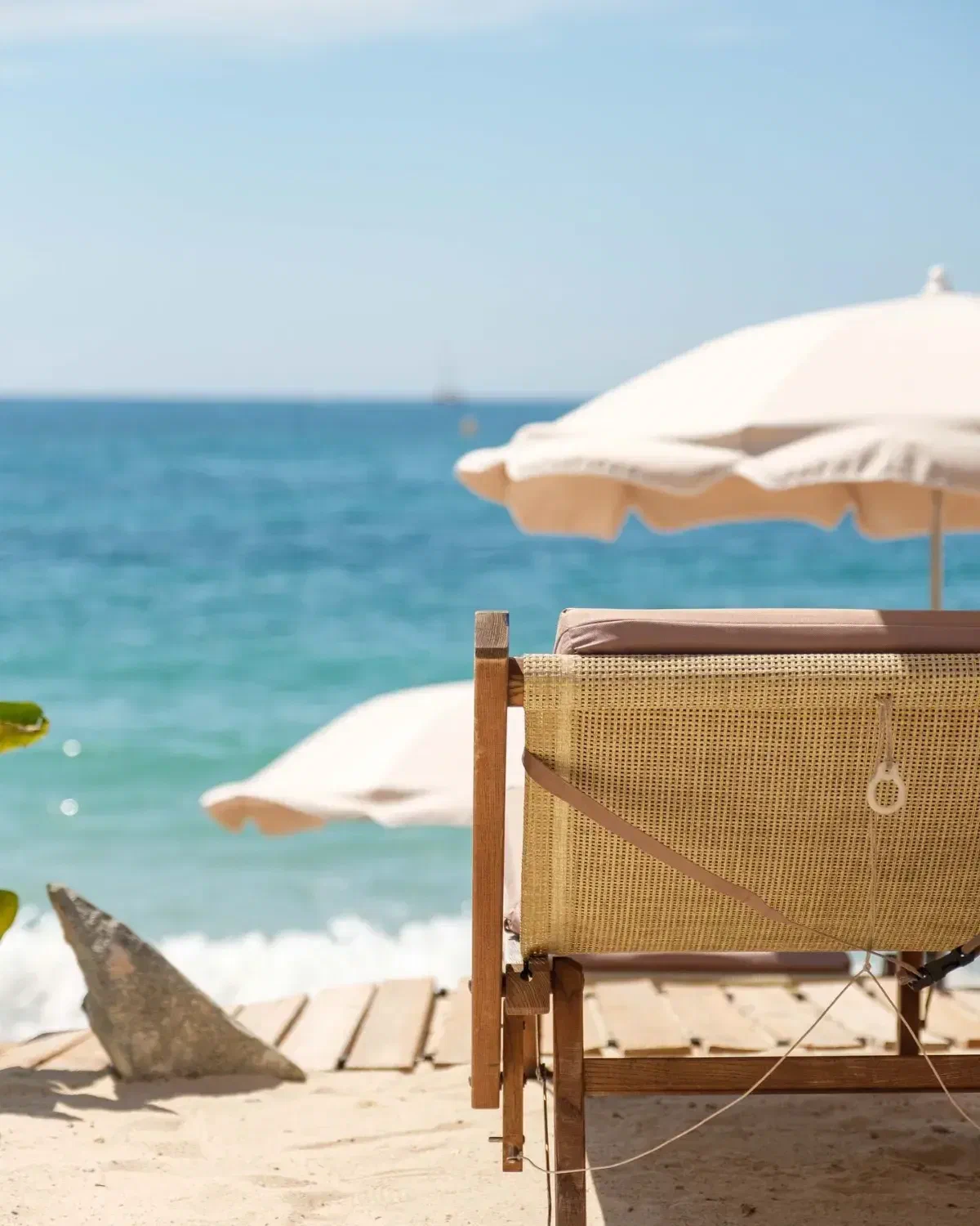 The back of an empty lounger on the sand, a cream parasol open above it and the sea behind.
