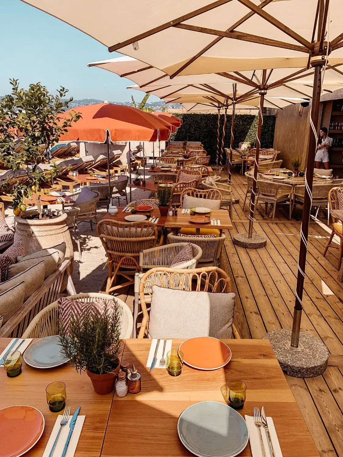 Tables laid under cream parasols late in the day, the light coming in low across the terrace.