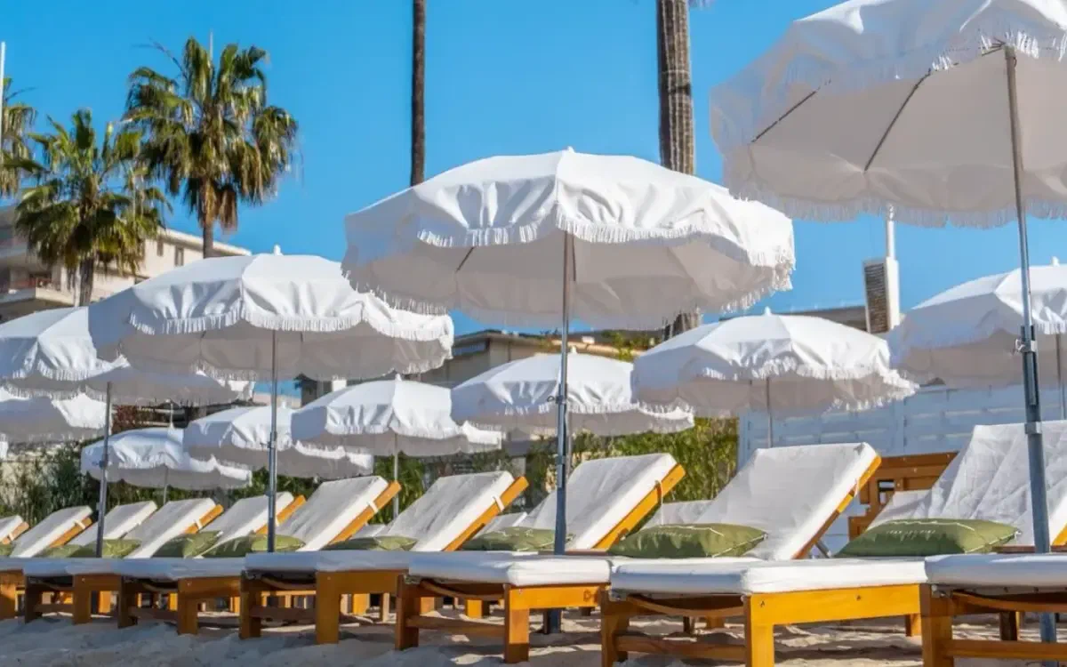 White fringed parasols standing over loungers on raked sand, palms above them against a clear sky.