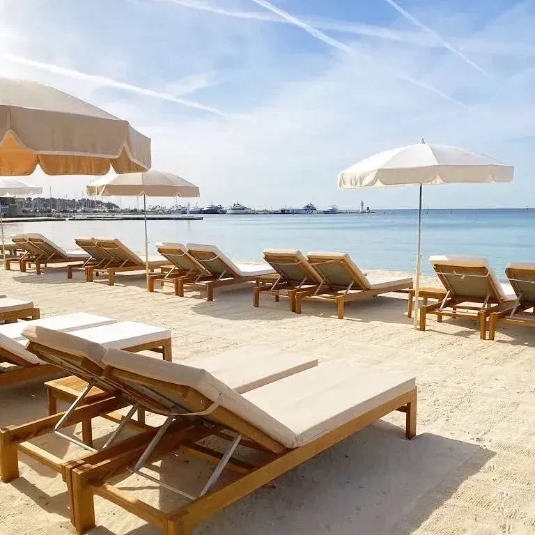 Loungers and pale parasols set out on raked sand, the marina and the water beyond them.