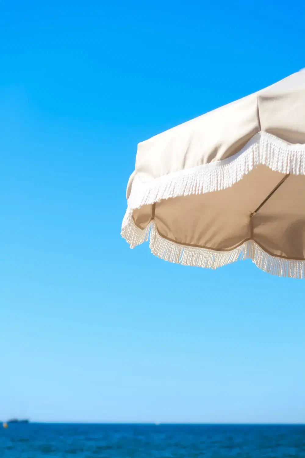 The edge of a cream fringed parasol against an open blue sky, the sea low beneath it.