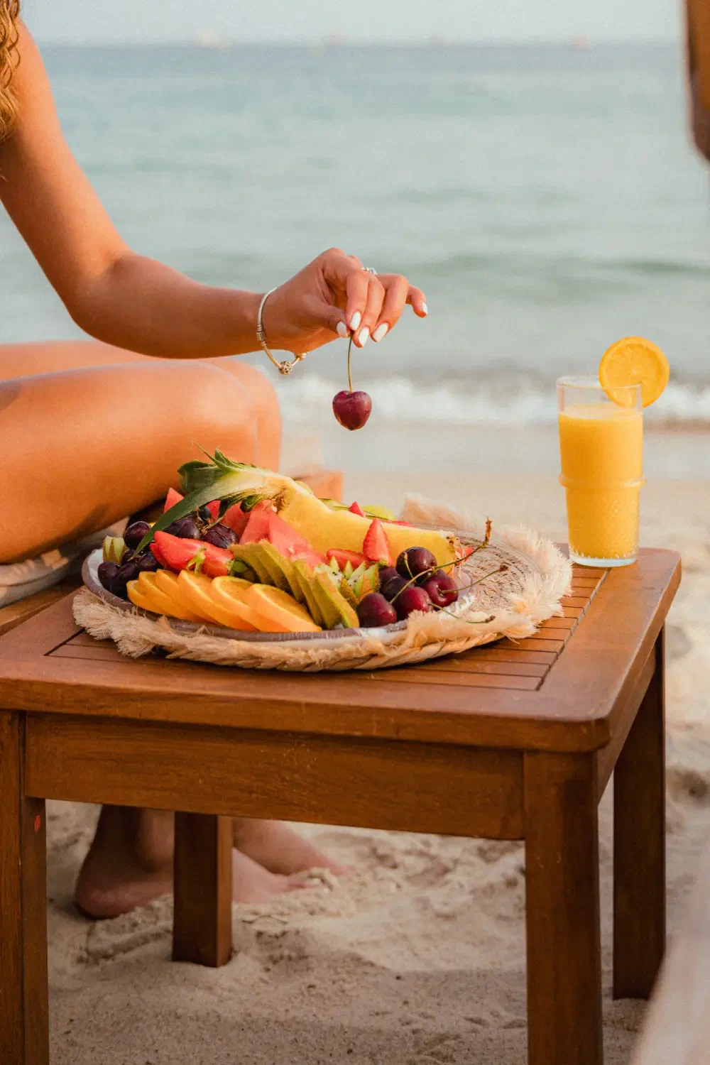 A hand lifting a cherry from a plate of fruit on a low teak table, the sea soft behind it.