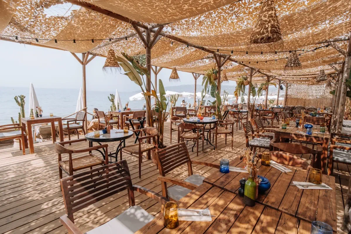 A beach restaurant under woven shade canopies, timber tables laid and the sea open at one side.
