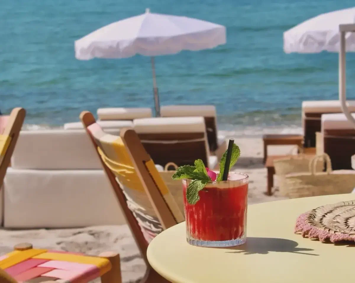 A tall red drink with mint on a round table above the sand, white parasols and the sea behind it.