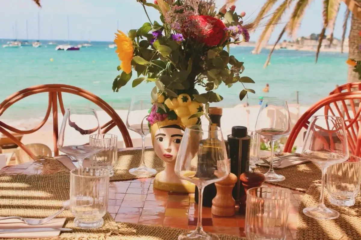 A table laid on a terrace above the water, flowers in the middle of it and boats out on the bay.