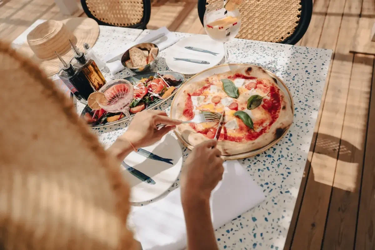 Seen from above, a table on a terrace with a pizza, a salad and two hands lifting a knife and fork.