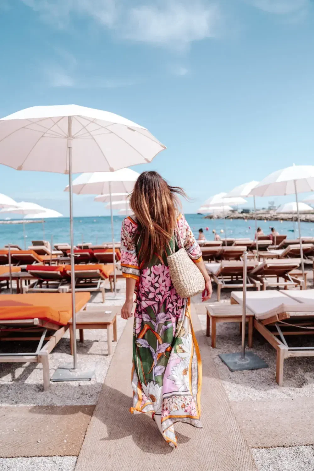 A woman with a straw basket walks down matting between rows of loungers towards the water.
