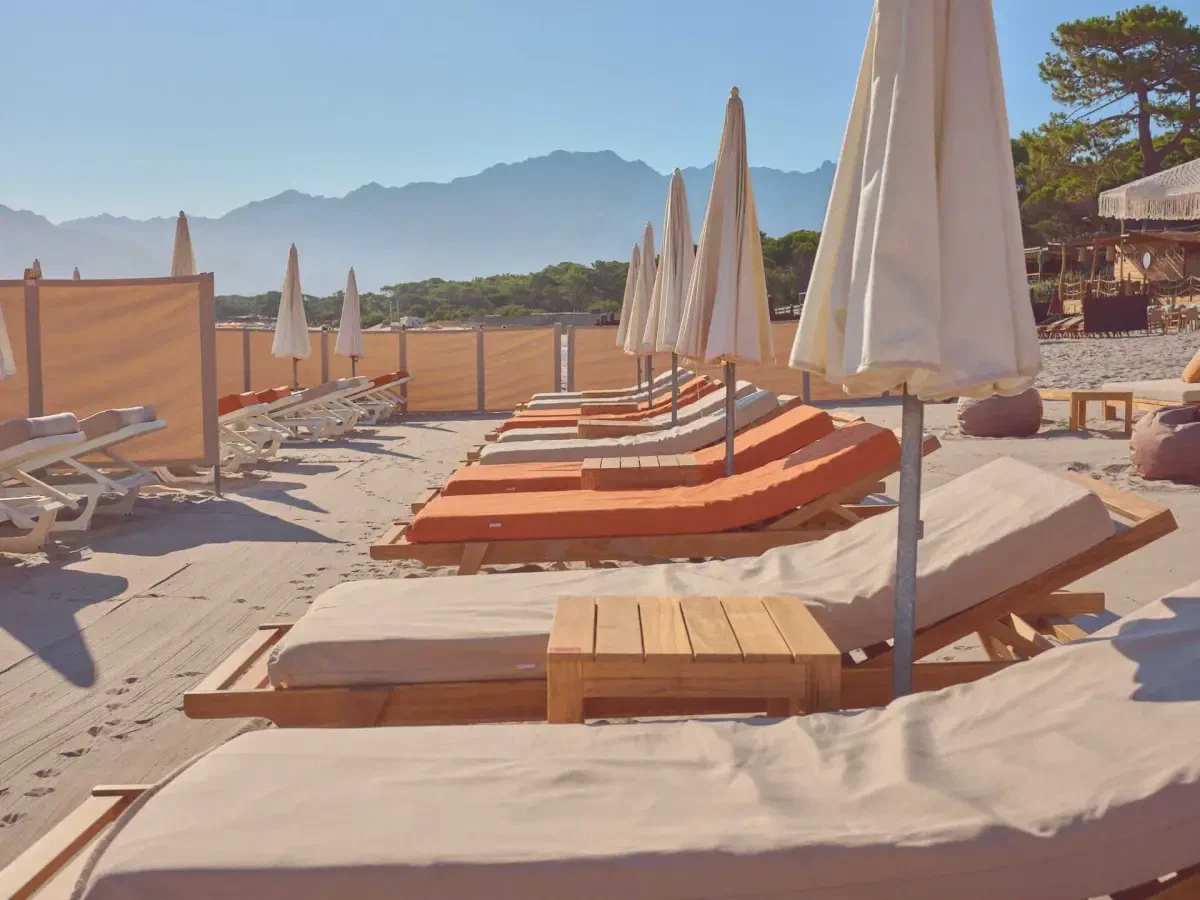 Loungers and folded parasols on pale sand late in the day, mountains standing behind in the haze.