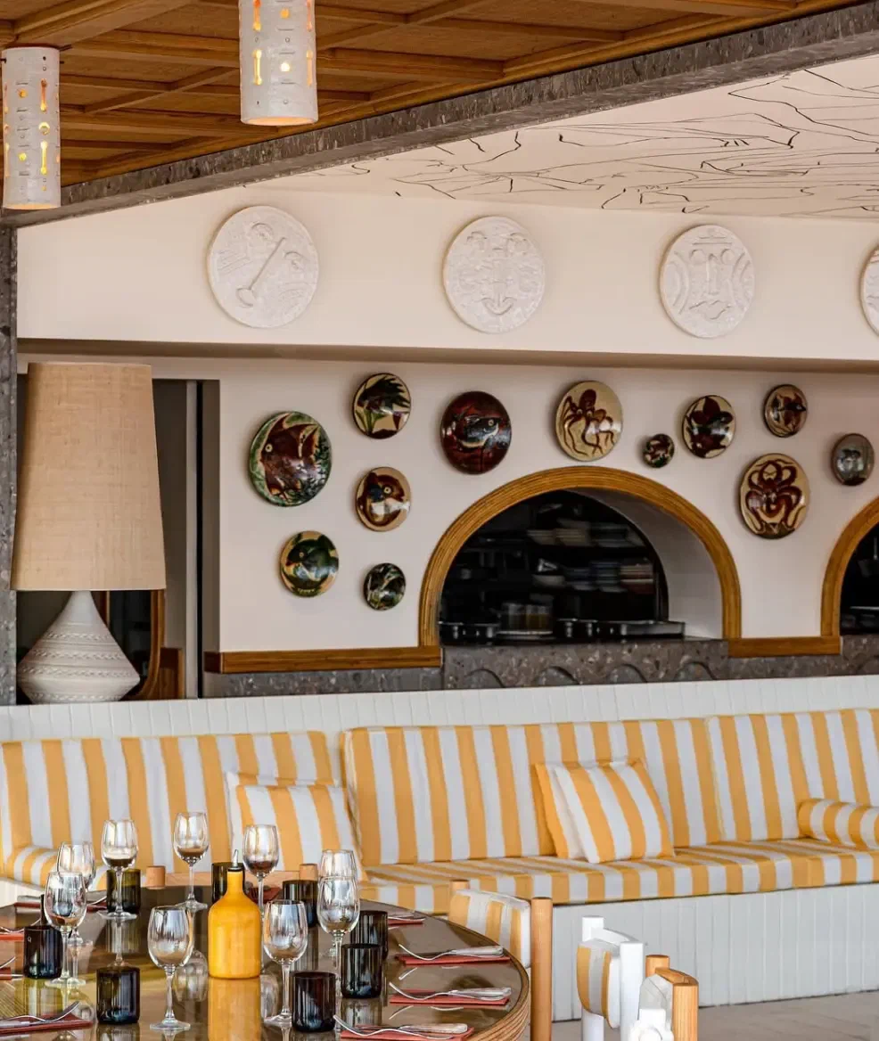 The inside of a beach restaurant, ceramic plates on a pale wall above a striped banquette and a laid table.